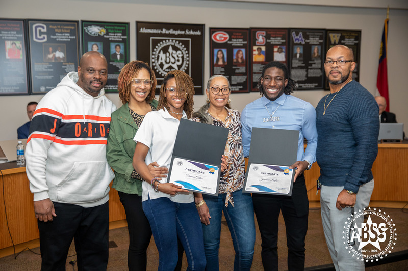  Group photograph of student athletes Jonathan Paylor and Deanna Cotton holding their certificates and standing with their family in front of the Board of Education