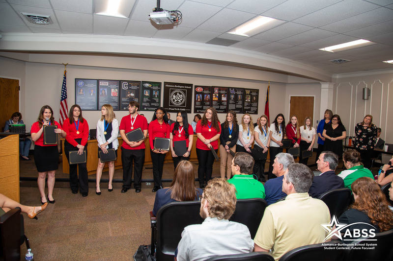 Photograph of a large group of Southern Alamance High School's FCCLA Club standing at the front of a board meeting and smiling or clapping as Board Member Donna Davis Westbrooks speaks with a microphone speaking to the audience