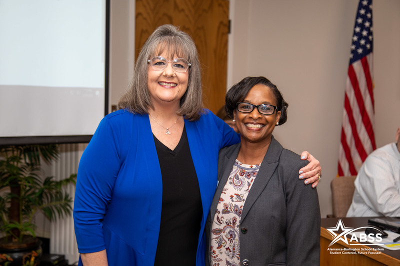 Village Project Executive Director standing with Board Member Donna Davis smiling in front of the audience at a board meeting