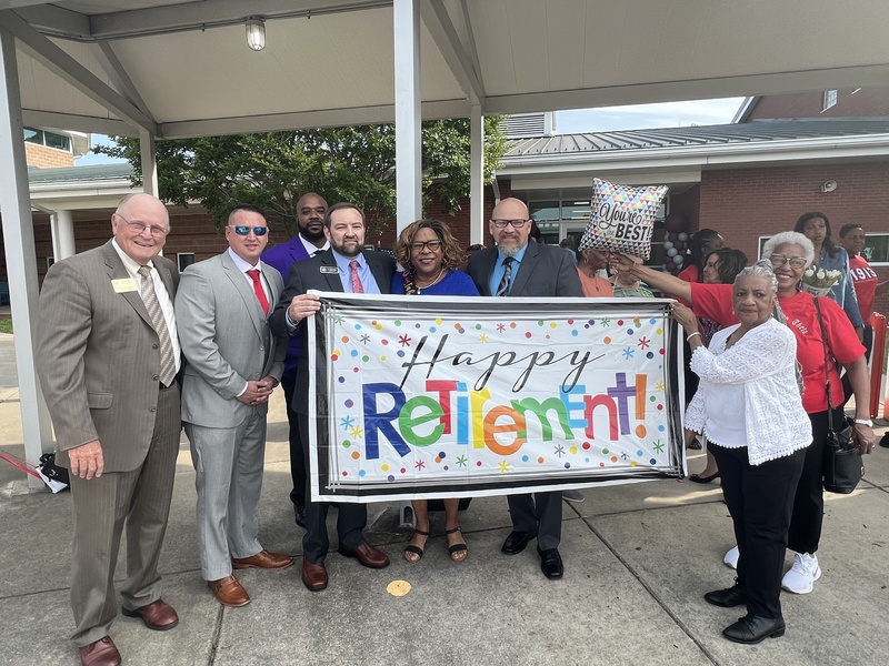 Dr. Butler and Board Members outside Highland Elementary with Patsy Simpson holding Happy Retirement Banner 