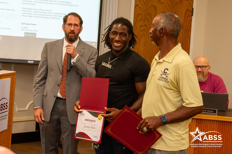 Jonathan Paylor stands with coaches while holding a certificate of recognition and Board member Dr. Charles Parker speaking behind him