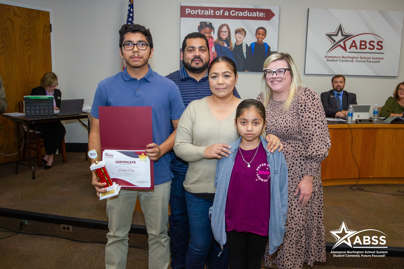 Alamance-Burlington Early College Student Hector Perez holding a STAR Student certificate and trophy standing with his family and principal Whitney Fleihman at a Board Meeting
